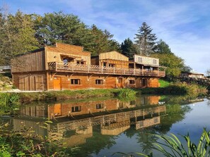 Een rustiek houten gebouw met balkon, gelegen aan een kalm waterlichaam, omringd door bomen.