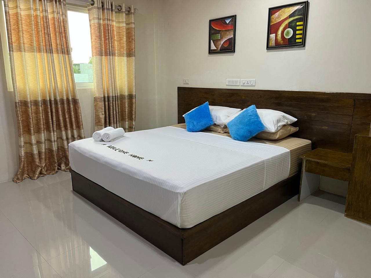 A hotel room with a large bed, a wooden headboard, two paintings on the wall, and a window with curtains.