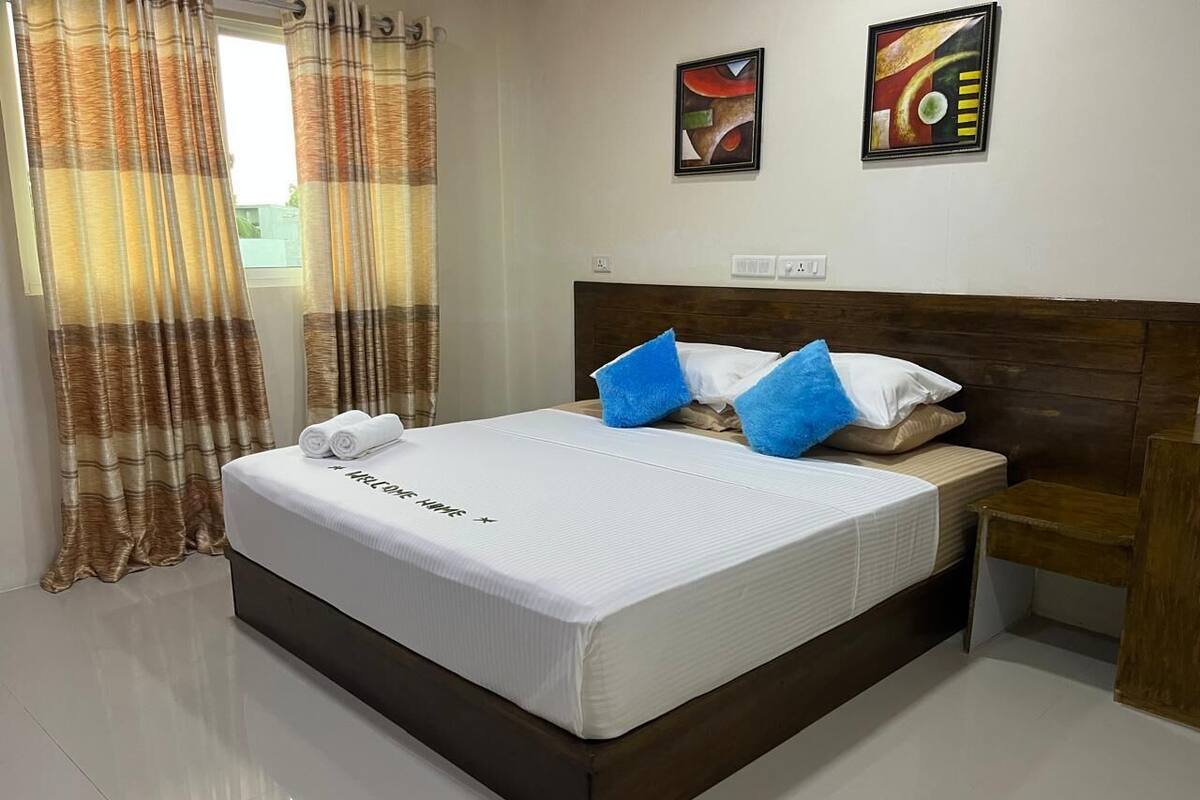 A hotel room with a large bed, a wooden headboard, two paintings on the wall, and a window with curtains.