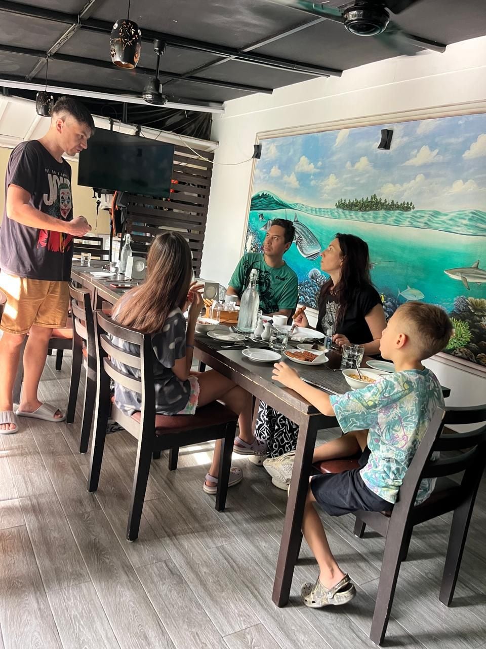 Zona pranzo con tavolo apparecchiato per sei persone, televisione a parete e un murale raffigurante una veduta costiera.