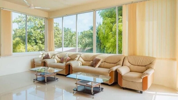 A spacious living room with a beige sofa set, two glass coffee tables, and large windows offering a view of greenery outside. - Oceanvine Maldives (Mahibadhoo)