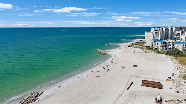 A beach with clear turquoise water, sandy shore, and a row of modern buildings with blue roofs.