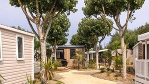 A pathway leading to a wooden cabin with a metal roof, surrounded by trees and a gravel area. - 24m² Mobile home with terrace in Saint-Jean-de-Monts (Saint-Jean-de-Monts)