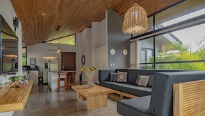 A modern living room with a wooden ceiling, a gray sofa, a wooden coffee table, and a large window with a view of greenery. - Villa Bernard - luxurious home in Tamarindo, walking distance to the beach& town (Tamarindo)