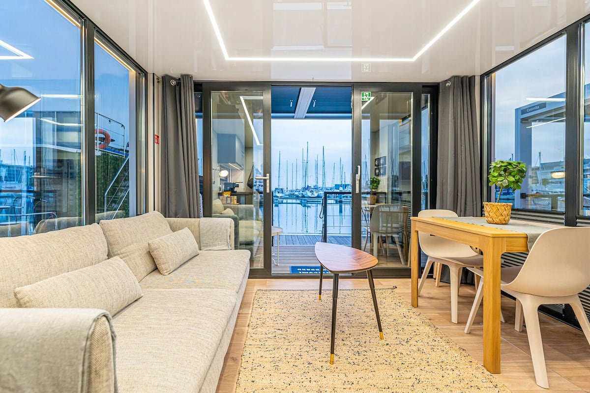 A modern living room with a sofa, a dining table, and chairs. There is a view of a marina through large glass doors.