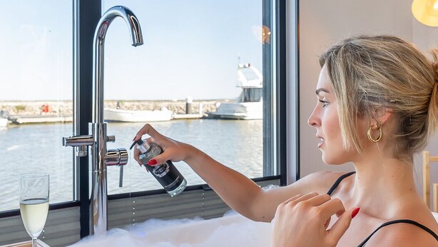 Una persona en una bañera con vistas a un muelle y barcos, una copa de vino y un bol de fruta.