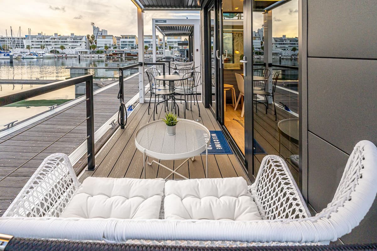 A deck with a white cushioned seating area, a small round table with a plant, and a view of the marina and buildings in the background.