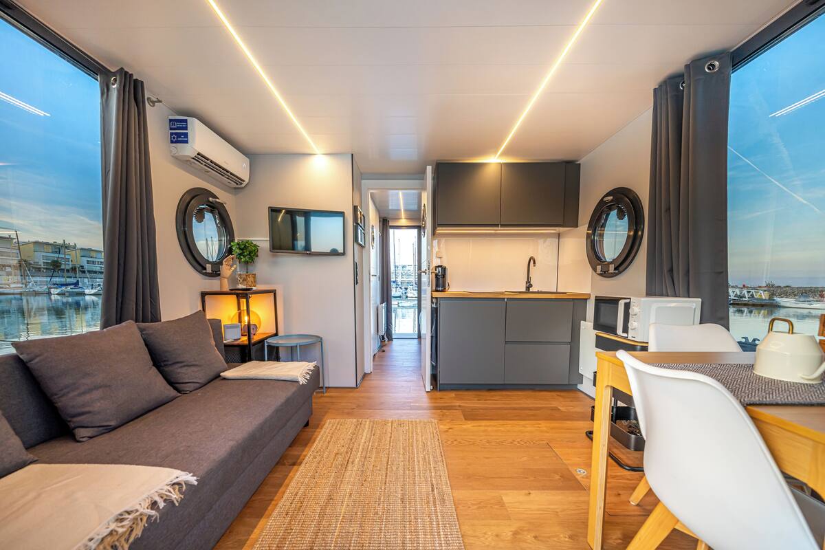 A modern, compact living space with a sofa, dining table, kitchen, and large windows overlooking a marina.