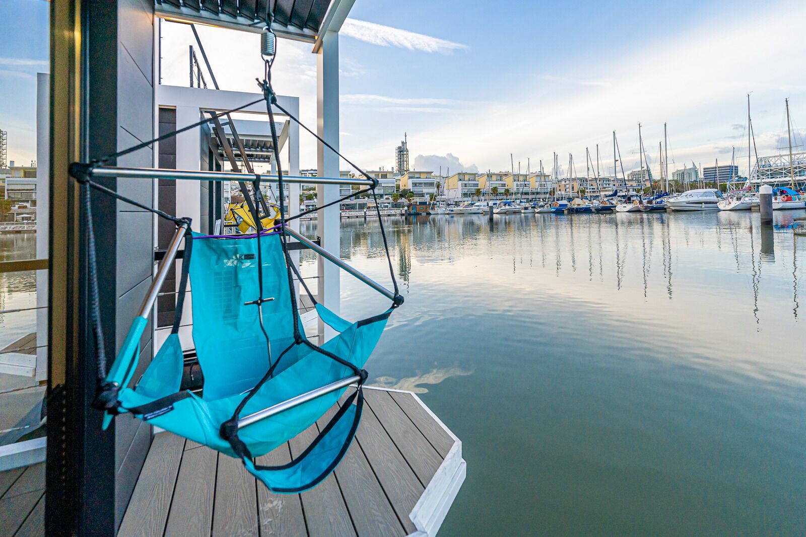 Uma cadeira suspensa azul sobre um deck de madeira, com vista para um porto com barcos e edifícios ao fundo.