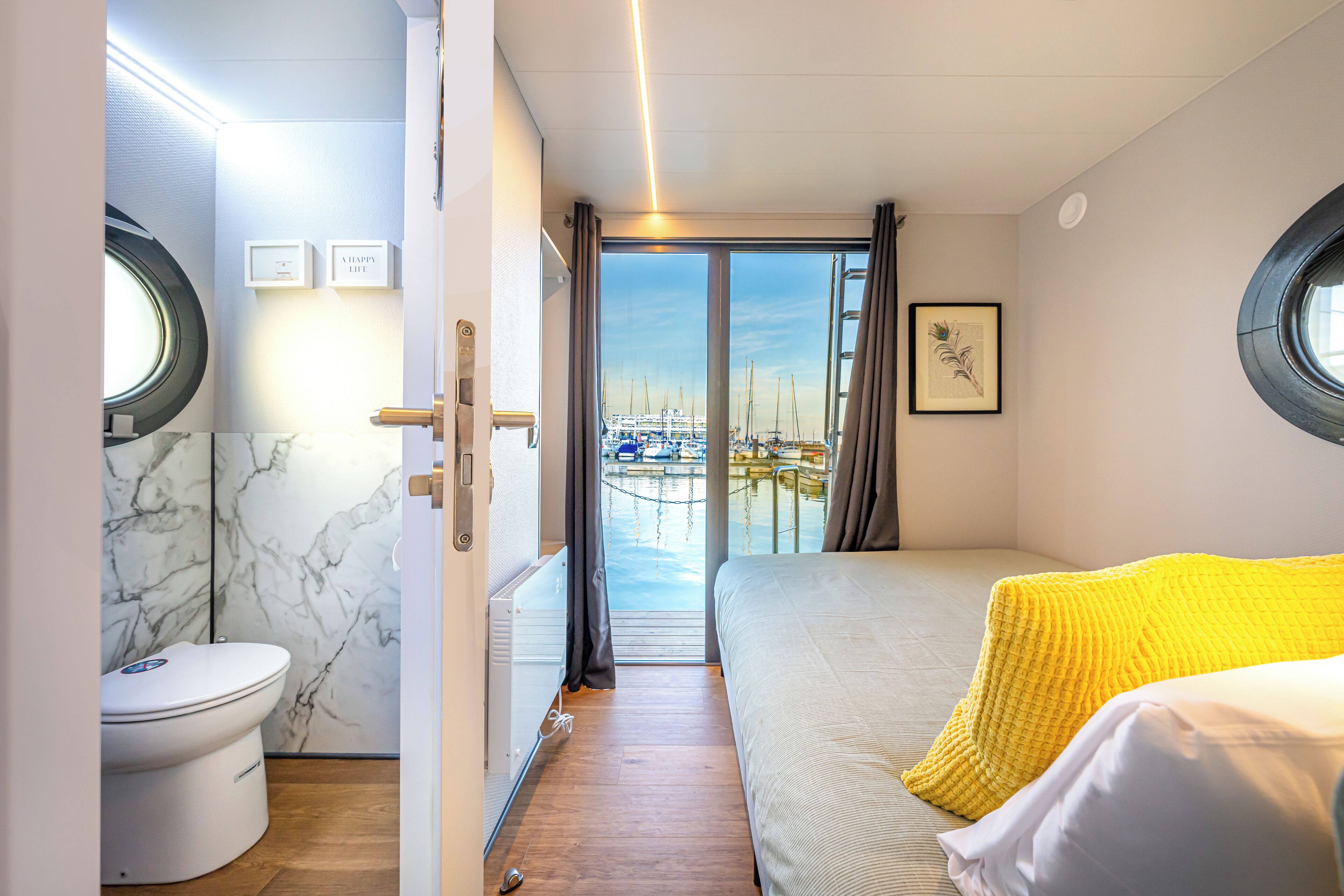 Une petite pièce comprenant un lit, des toilettes et donnant sur une marina grâce à une porte vitrée.