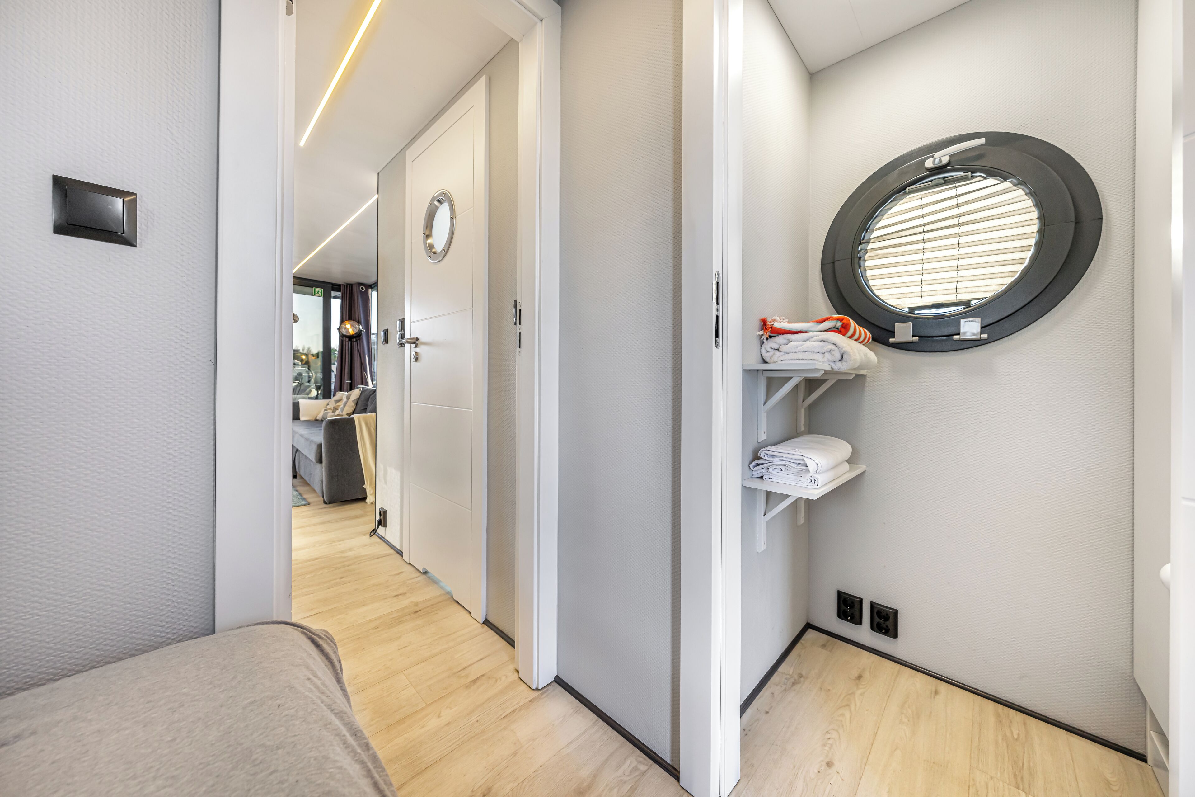 Un couloir étroit, avec un lit sur la gauche, une porte au fond et une fenêtre ronde avec des stores sur la droite.