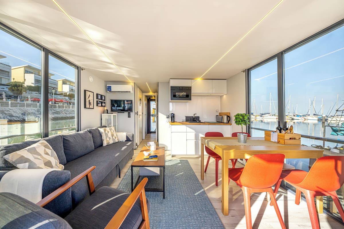 A modern living room with a sofa, coffee table, and dining area with chairs. A kitchen is visible in the background with a view of the marina through large windows.