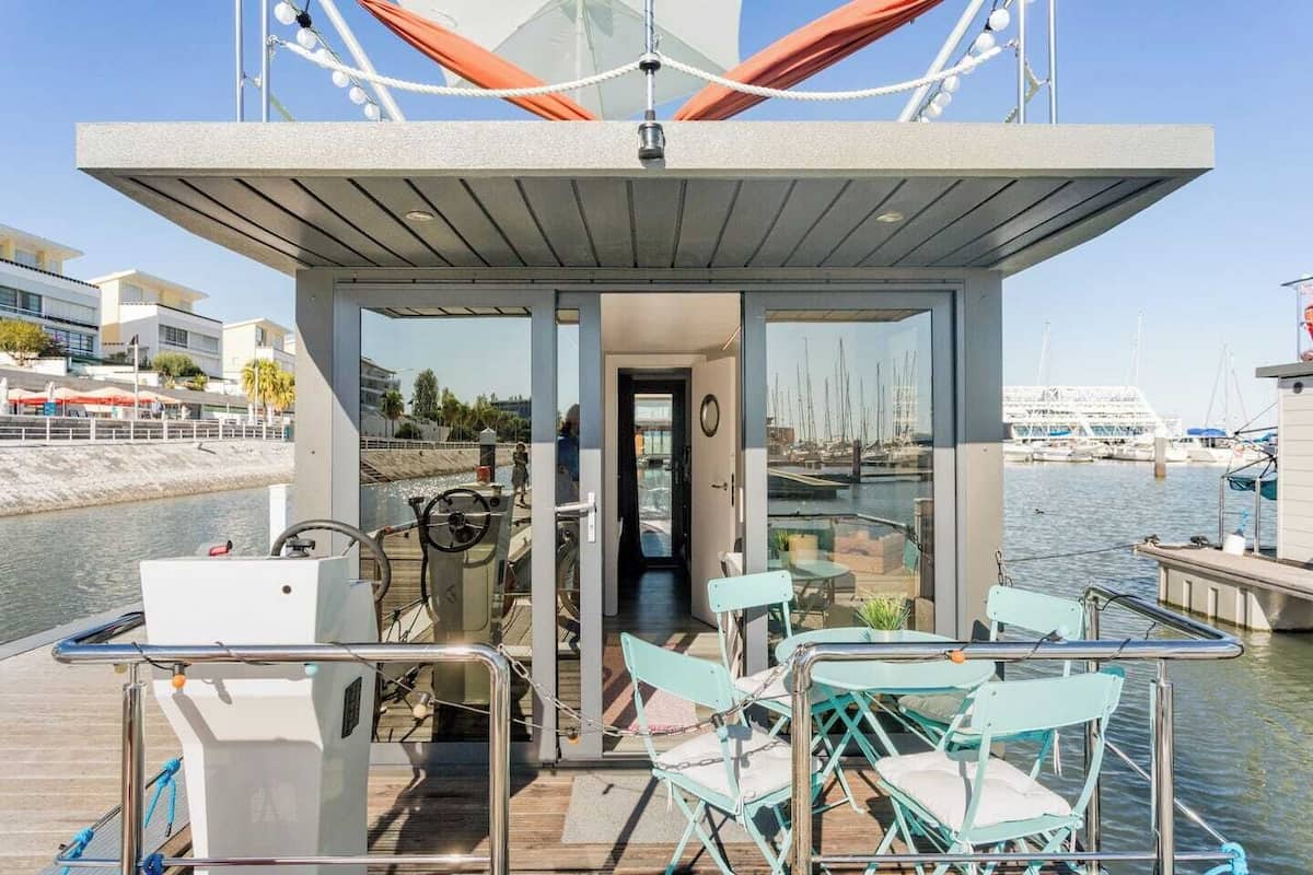 A small, enclosed deck area with a table and chairs, a steering wheel, and a view of the marina through glass doors.