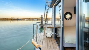 Un barco moderno con zona de descanso cubierta de vidrio, cubierta de madera y vista al agua y edificios distantes.