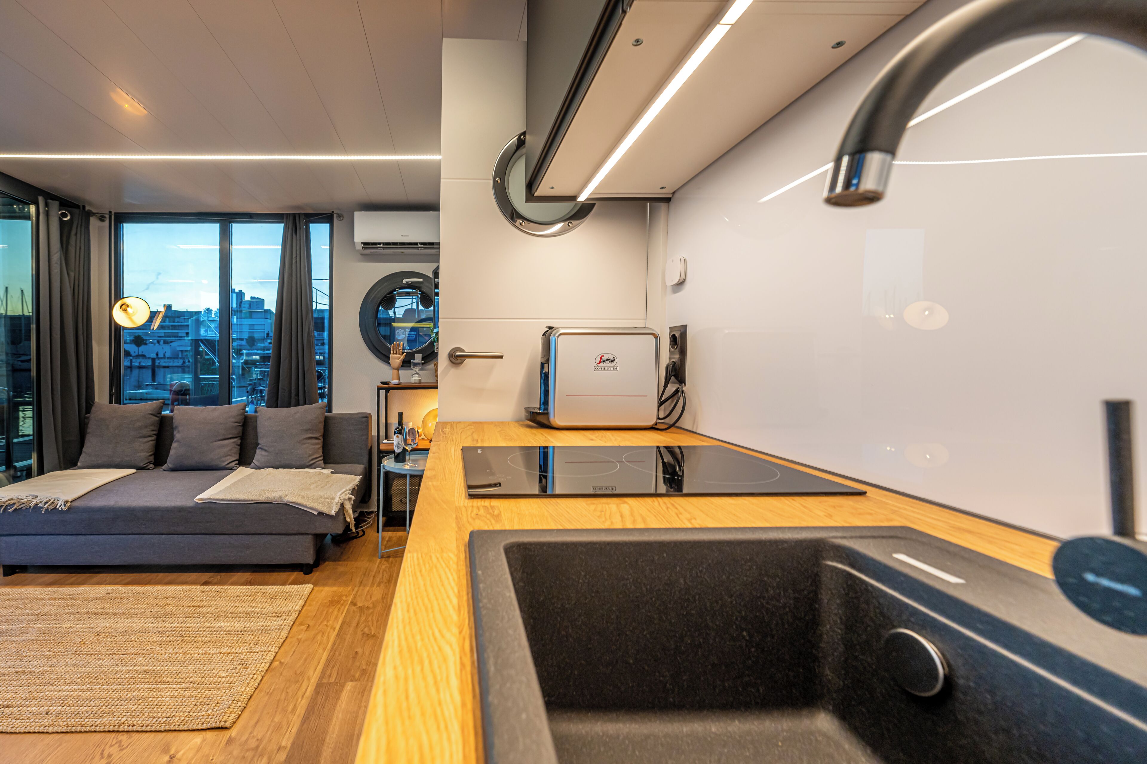 Un salon moderne avec un canapé, un petit coin cuisine doté d’un évier et une vue sur la ville grâce à de grandes fenêtres.