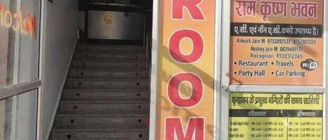 A hotel entrance with a staircase leading to the rooms. There are posters on the walls, including one with the word "ROOM" and another with a list of amenities like a restaurant and car parking.