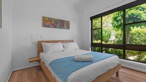 Une chambre avec un lit en bois, du linge de lit blanc, un plaid bleu, un tableau au mur et une grande fenêtre donnant sur un paysage verdoyant.