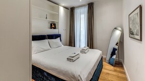 A neatly made bed with white linens and two towels, a bedside mirror, a shelf with a small item, and a window with curtains. - Sabina 2 -bedroom apartment with kitchen in center Roma (Roma)