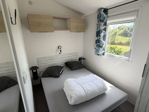 A single bed with a headboard, two bedside tables, a window with curtains, and built-in wooden shelves above the bed. - Mobile home 40m², 4 rooms, equipped kitchen (Binic)