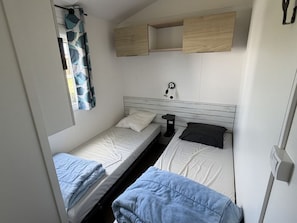 Ein kleines Zimmer mit zwei Betten, einem kleinen Tisch, einer Wandlampe und einem Fenster mit Vorhang.