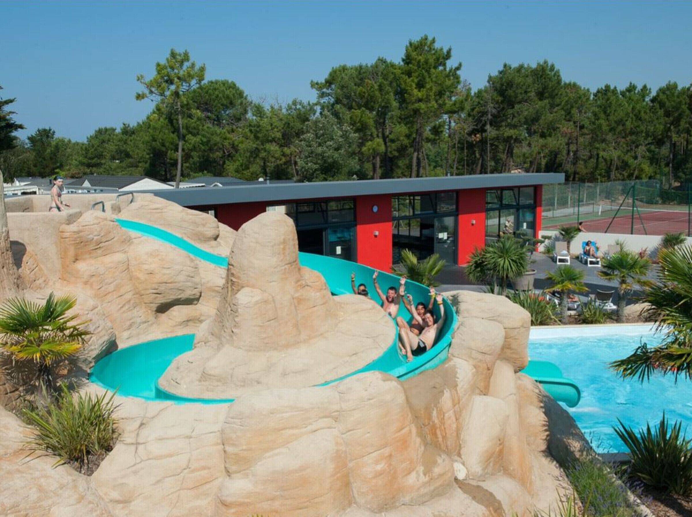 A water slide with a rocky design, a pool, and a modern building with red and grey architecture.