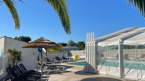 Ett poolområde med solstolar, en möblerad sittgrupp under tak och en pool med en genomskinlig avskärmning.