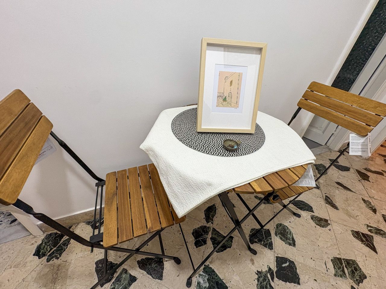 Una pequeña mesa con un cuadro enmarco, dos sillas de madera y un suelo estampado.