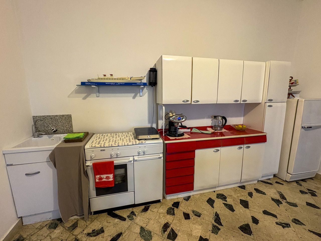 Une cuisine de taille réduite avec des meubles blancs, un plan de travail rouge, une cuisinière, un évier et un petit réfrigérateur.