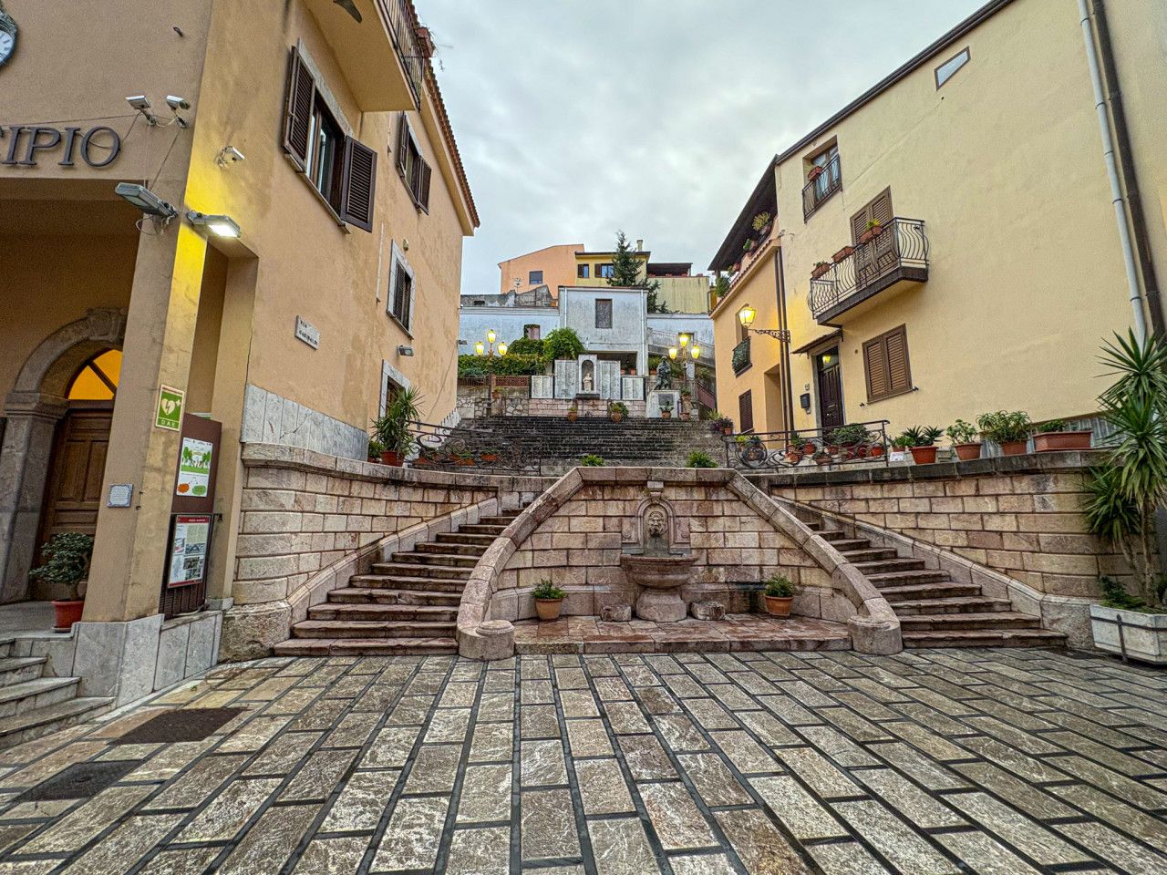 Eine Steintreppe führt zu einem Gebäudeeingang, der von einem Brunnen und Kübelpflanzen gesäumt ist.