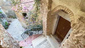 Una escalera de piedra que lleva a una puerta de madera, rodeada de un muro de piedra rústico y un pequeño jardín con macetas de plantas y flores.