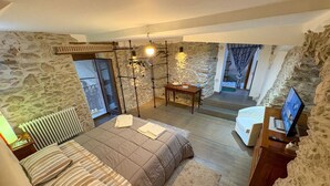 Un dormitorio con una cama grande, una mesita de noche de madera, un escritorio con una computadora y una pared de piedra. Hay una ventana con cortinas y vistas al exterior.