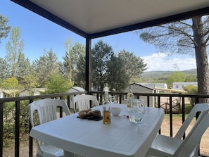 A covered outdoor area with a table set for two, surrounded by white chairs, and a view of trees and buildings in the background. - Mobile home with terrace in Villecroze (Villecroze)