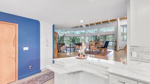 A modern kitchen with white cabinetry and a marble island, a dining area with a table and chairs, and a view of the outdoors through large windows.