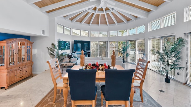 A spacious dining area with a high ceiling and large windows, a wooden cabinet, and a television in the background.