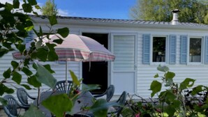 Een witte stacaravan met blauwe luiken, een terras met stoelen en een gestreepte parasol.