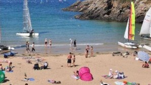 Een strand met mensen die zonnebaden en zwemmen, boten op het water en rotsachtige kliffen op de achtergrond.