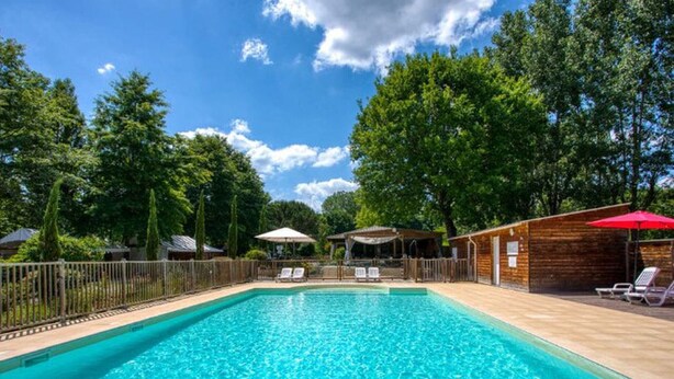 A rectangular swimming pool with clear blue water, surrounded by a wooden deck, and shaded by trees and umbrellas. - 27m² Mobile Home with Terrace in Groléjac (Groléjac)