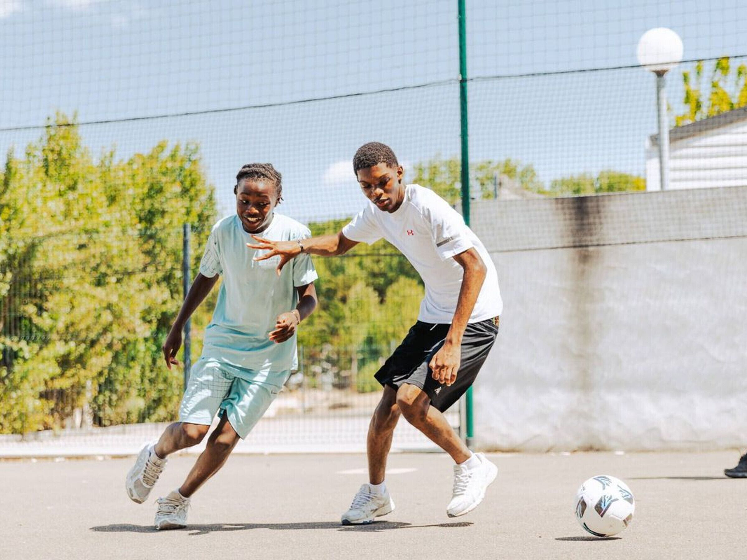 Zwei Personen spielen Fußball auf einem Außenplatz mit Netz und Bäumen im Hintergrund.