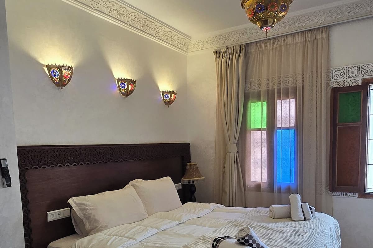 A hotel room with a large bed, decorative wall sconces, a patterned rug, and a remote control on a small table.