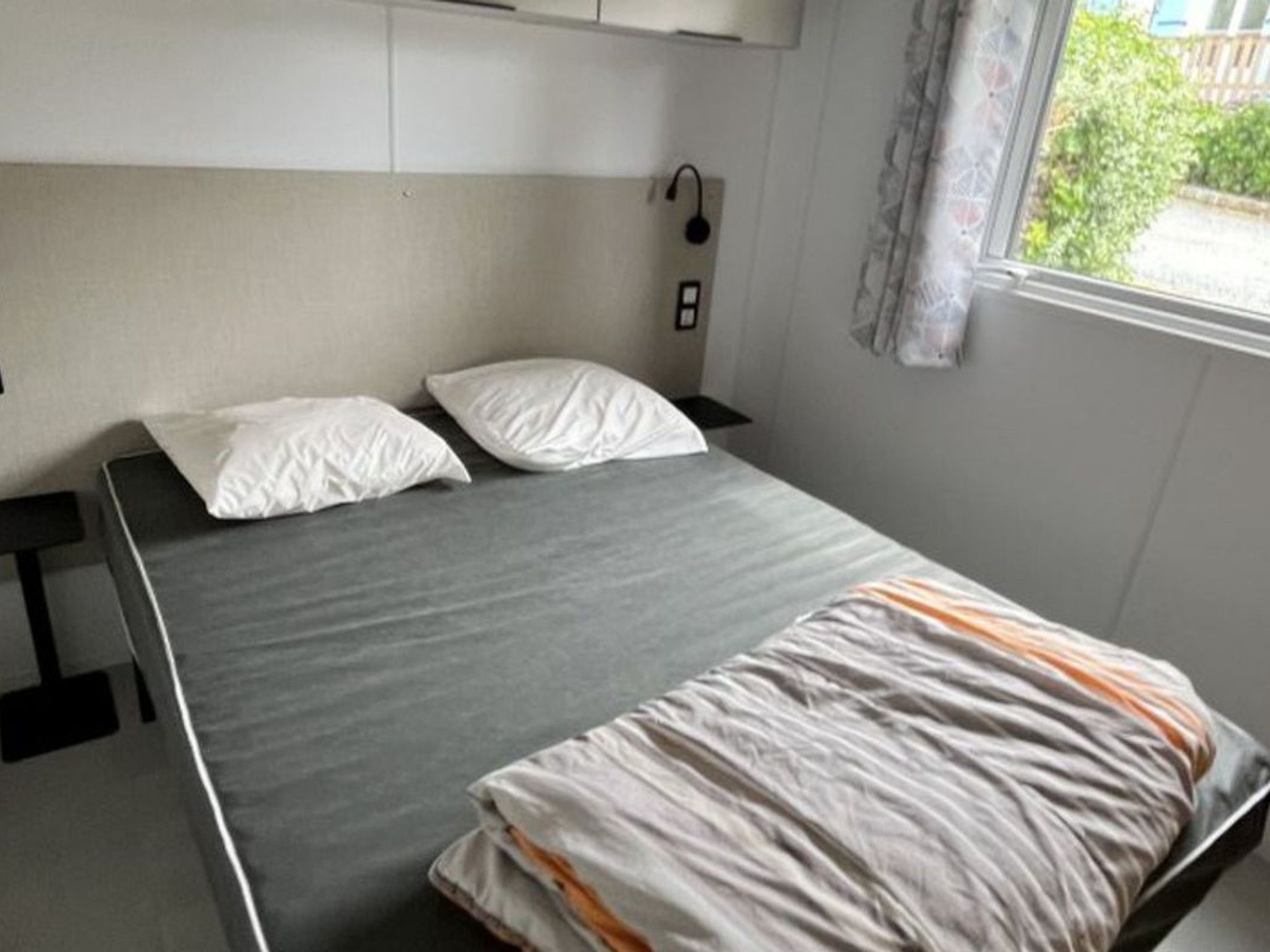 Un letto singolo con materasso grigio e cuscini bianchi, un comodino e una finestra con tenda.