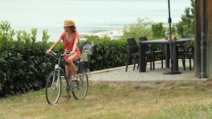 Una persona in bicicletta vicino a un tavolo e delle sedie, con vista su un lago e vegetazione sullo sfondo.