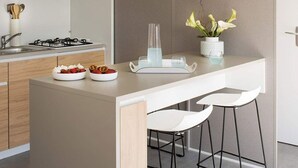 Una cocina moderna con una isla central, dos taburetes blancos de bar, una estufa a gas y un pequeño estante con adornos.