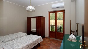 Una stanza con due letti, un armadio in legno, un tavolo con una ciotola decorativa, un lampadario a sospensione e una porta scorrevole in vetro che dà su una zona esterna.