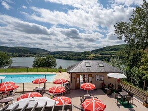 Ein Poolbereich mit roten Sonnenschirmen, Sitzgelegenheiten im Freien und Blick auf einen See und Hügel.