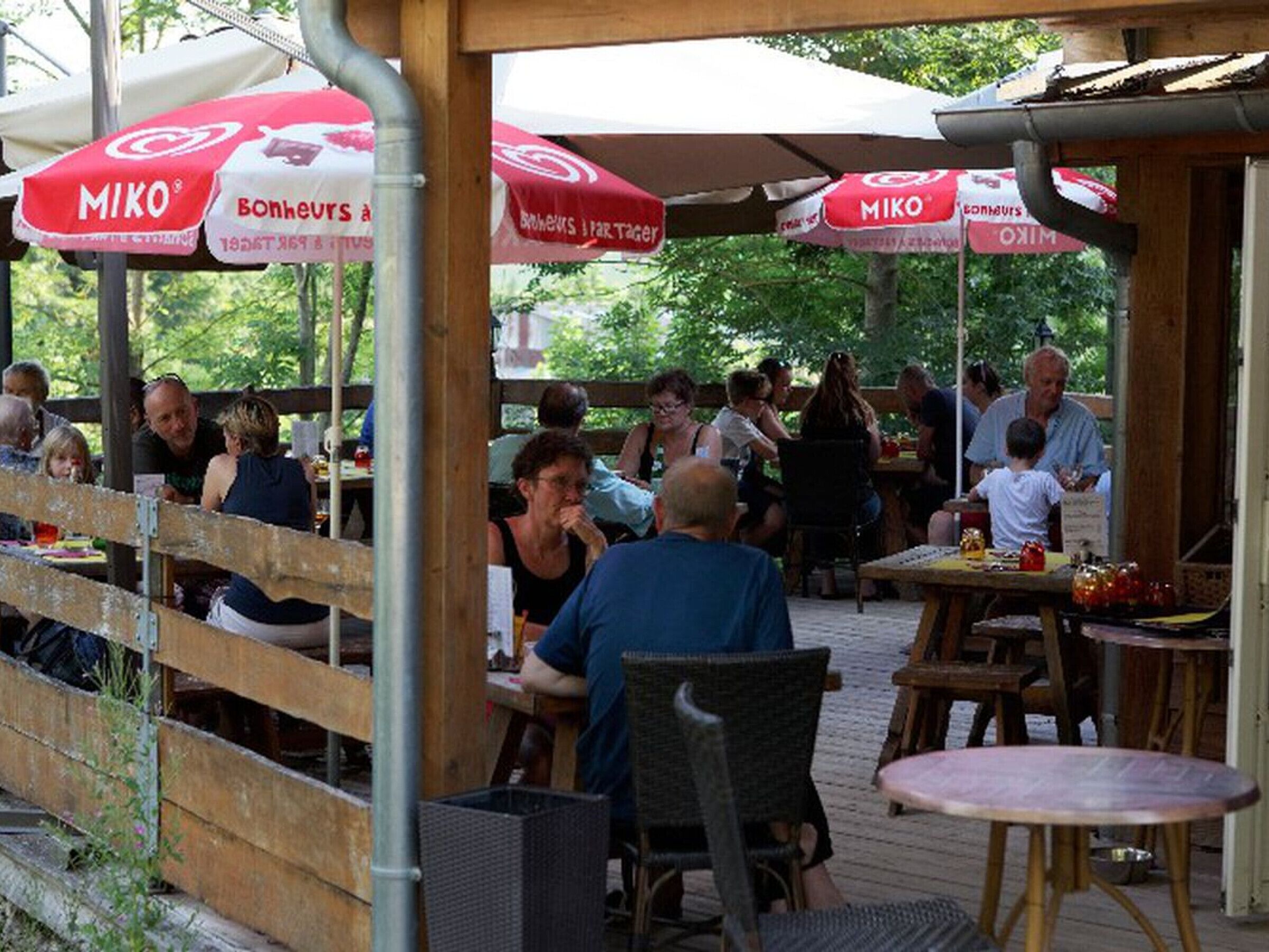 An outdoor dining area with wooden tables and chairs, covered by red and white umbrellas with 'MIKO' branding.