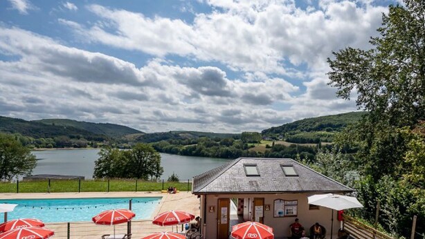 Een zwembadterras met rode parasols, buitenmeubilair en uitzicht op een meer en heuvels.