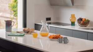 Une cuisine moderne avec un plan de travail blanc, une miche de pain, un pichet en verre, un saladier de fruits et une machine à café.