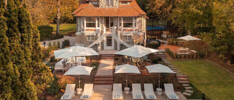 A large house with a swimming pool, surrounded by lounge chairs and umbrellas.