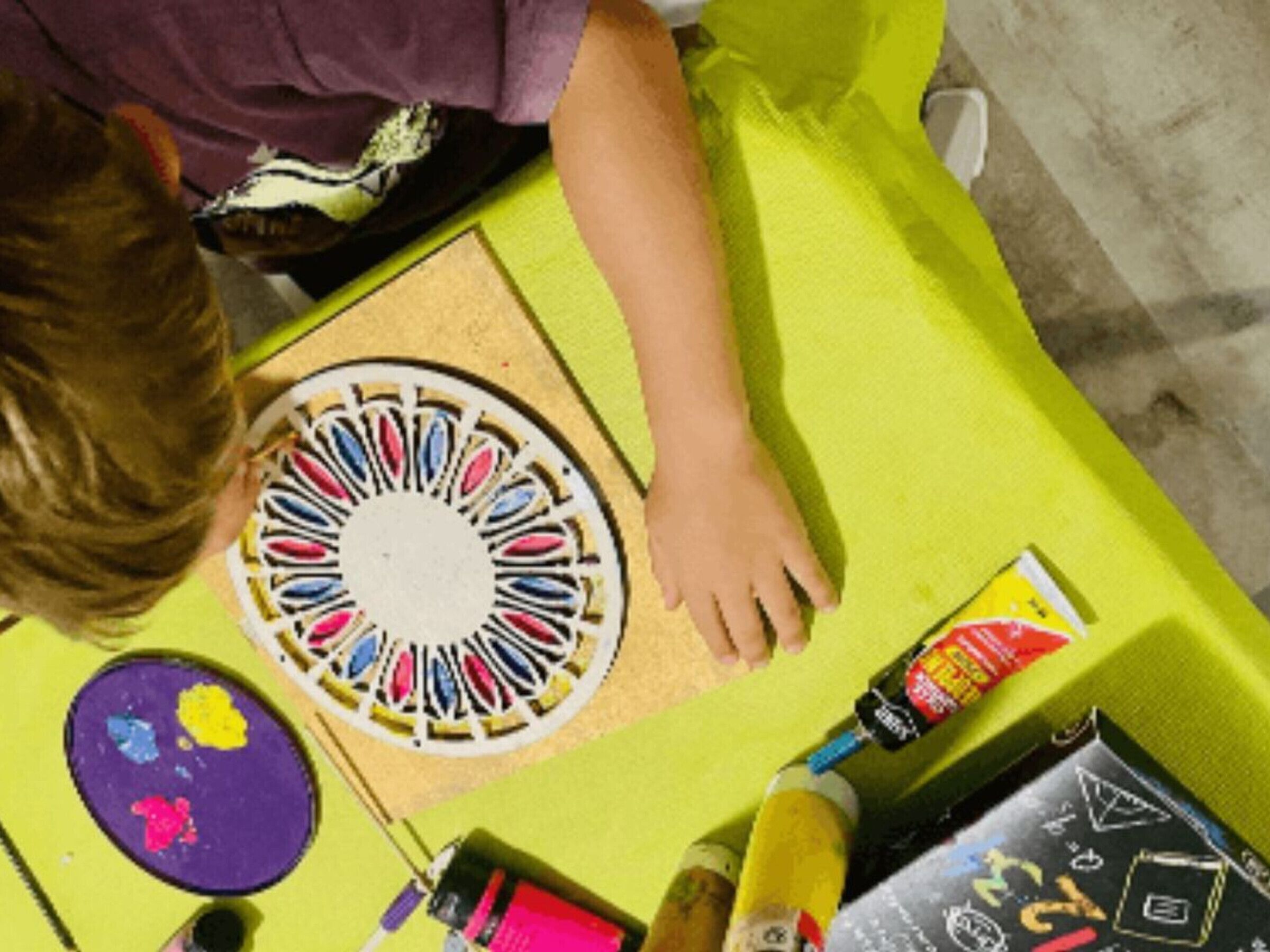 Un enfant est en train de peindre sur une toile ronde, avec des pinceaux et des tubes de peinture à proximité.