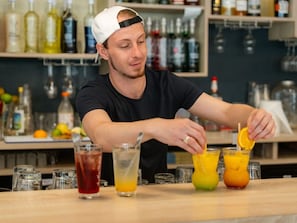 Un barman prépare des boissons dans un bar, avec diverses bouteilles et différents verres en arrière-plan.